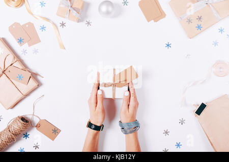 Belle femme mains tenant une petite boîte blanche avec un cadeau sur fond blanc parmi les décorations de Noël, vue du dessus Banque D'Images