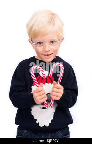 Noël d'amour : un mignon petit garçon de 4 ans portant des lunettes et un pull-over de père noël tient deux bâtons bonbons, formant une forme de coeur sur un blanc parfait Banque D'Images