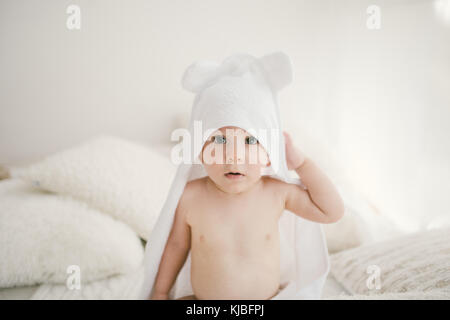 beau bébé garçon souriant recouvert d'une serviette blanche avec des oreilles amusantes. Assise sur un intérieur en laine à carreaux blanc. Banque D'Images