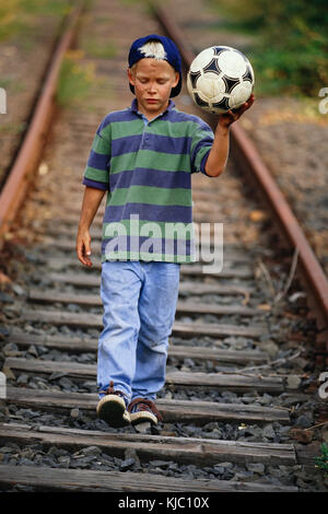 Garçon avec un ballon de football marchant le long des voies de train Banque D'Images