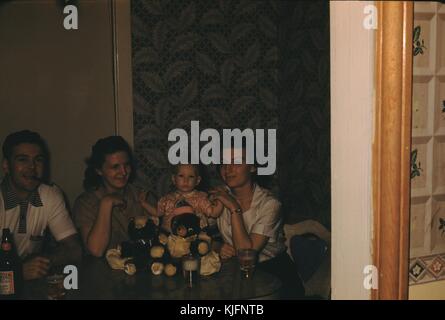 Une photographie de trois adultes et un bébé assis à une table, il y a trois verres de bière sur la table, l'homme assis à la table fume un cigare et a une bouteille de bière devant lui, l'enfant est entre les deux femmes et a deux ours en peluche devant elle, les adultes sourient tous pour le photographe, 1952. Banque D'Images