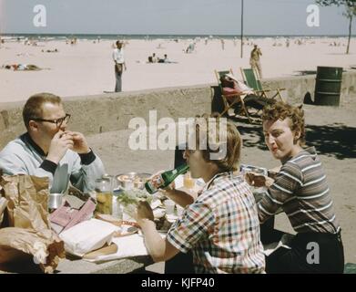 Trois personnes, un homme et deux femmes, assis à une table, sur une promenade en béton à côté de la plage, mangeant une grande quantité de nourriture et buvant une boisson gazeuse, en arrière-plan, des gens allongés sur le sable à la plage, 1952. Banque D'Images