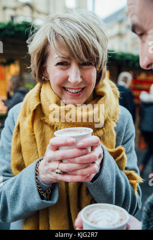 Mature femme sourit à l'appareil photo tout en buvant un chocolat chaud dans le marché de Noël. Banque D'Images