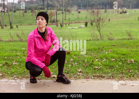 Jeune femme l'analyse de la piste avant l'exécution par une froide journée d'hiver sur la voie de la formation d'un parc urbain. athlète féminin portant un coupe-vent rose, b Banque D'Images