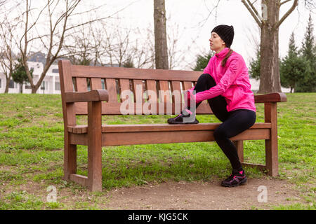 Femme athlète fatigué ou déprimé se reposant sur un banc sur une froide journée d'hiver sur la voie de la formation d'un parc urbain. Jeune femme portant un coupe-vent rose Banque D'Images