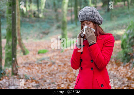 Jeune femme souffrant de rhume ou grippe soufflant du nez ou de l'éternuement sur du papier blanc mouchoir en forêt portant un long manteau ou pardessus, un beanie un Banque D'Images