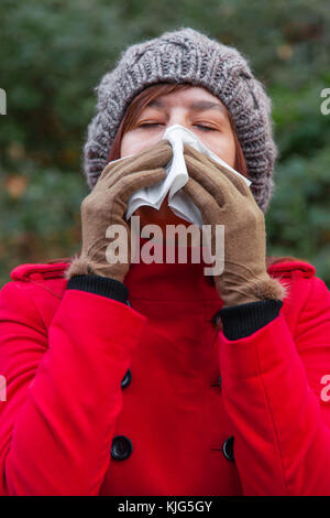 Jeune femme souffrant de rhume ou grippe soufflant du nez ou de l'éternuement sur du papier blanc mouchoir en forêt portant un long manteau ou pardessus, un beanie un Banque D'Images