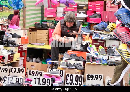 Vieux stallholder sur Dalston Market regardant le téléphone sur le stand de chaussures Banque D'Images
