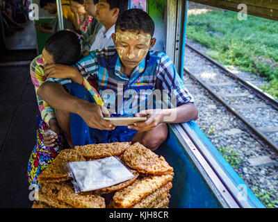 Yangon, région de Yangon, Myanmar. 25 novembre 2017. Un vendeur de snacks voyageant avec sa femme vérifie son téléphone intelligent dans le train circulaire de Yangon. Le Yangon Circular train est un système de boucle à deux voies de 45 kilomètres (28,5 mi) de 39 stations qui relie les villes satellites et les banlieues au centre-ville. Le train a été construit pendant la période coloniale britannique, la deuxième voie a été construite en 1954. Les trains circulent actuellement dans les deux sens (sens horaire et sens antihoraire) autour de la ville. Les trains sont le moyen le moins cher de traverser Yangon et ils sont très populaires auprès de la classe ouvrière de Yangon. Vers 100 000 Banque D'Images