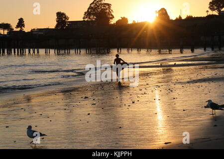 Silhouette d'un jeune homme portant sa planche sur la plage au coucher du soleil, Capitola, CA. Banque D'Images