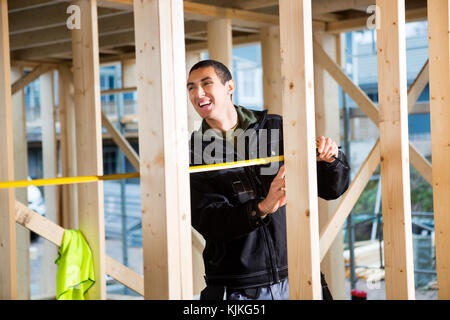 Young male carpenter rire pendant la mesure de bois at construction site Banque D'Images