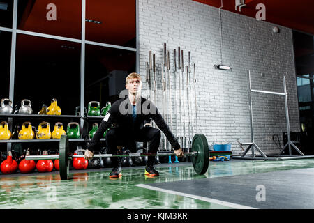 Le mec soulève le bar dans la salle de sport sur l'arrière-plan d'une fenêtre et un mur de brique gris Banque D'Images