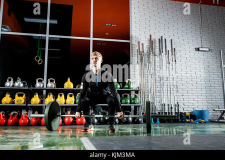 Le mec soulève le bar dans la salle de sport sur l'arrière-plan d'une fenêtre et un mur de brique gris Banque D'Images