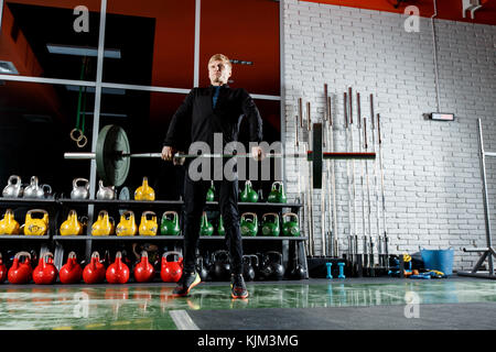 Le mec soulève le bar dans la salle de sport sur l'arrière-plan d'une fenêtre et un mur de brique gris Banque D'Images