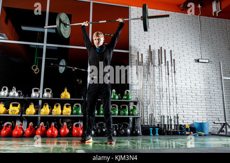 Le mec soulève le bar dans la salle de sport sur l'arrière-plan d'une fenêtre et un mur de brique gris Banque D'Images