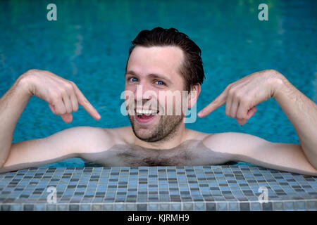 Fier jeune homme de race blanche se réjouit montrant avec index sur lui-même. Il est la natation en piscine durant les vacances Banque D'Images