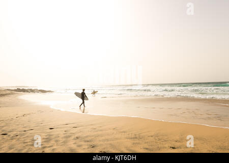 Deux surfeurs mâles dans leurs combinaisons de marche de la mer, les vagues sur une plage vide avec leurs planches de surf au coucher du soleil Banque D'Images
