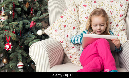 Cute preschool girl écrit lettre au Père sur tablette numérique, assis en face de l'arbre de Noël Banque D'Images