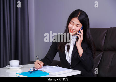 Jeune femme en costume d'affaires parler sur un téléphone mobile et rédige une note sur l'ordinateur portable Banque D'Images