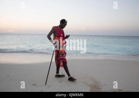Zanzibar, Tanzanie ; jeune guerrier Massaï sur la plage pour vendre bijoux Massaï et de l'artisanat traditionnel de leur cage à proximité pour les touristes à la plage hôtels vérifie son téléphone mobile. Banque D'Images