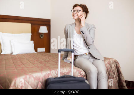 Portrait de jeune femme parlant au téléphone et smiling while sitting on bed in hotel room arrivant pour les voyages d'affaires, copy space Banque D'Images