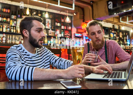 Portrait de deux hommes modernes using laptop sitting at table in bar travailler et profiter de la bière Banque D'Images