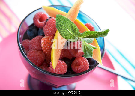 Fraise,framboise, myrtille et mangue salade de fruits frais garnie avec de la menthe fraîche dans un bol en verre coloré sur tapis de table. Banque D'Images