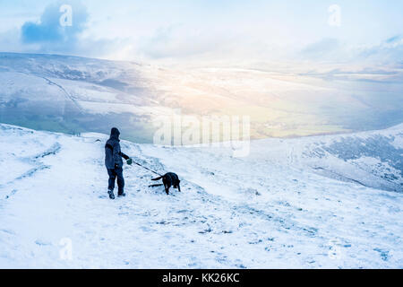 Promenade de chiens dans la neige. Randonneur avec chien randonnées avec la lumière du soleil qui brille sur une colline dans la vallée. Grand Ridge, Peak District, Derbyshire, Angleterre, RU Banque D'Images