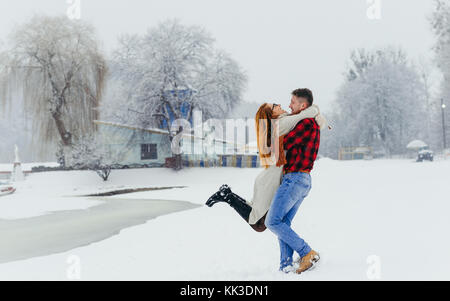 Heureux couple aimant s'amuse dans la forêt d'hiver. l'homme est en train de tourner autour de la belle femme tête rouge au cours de la neige. Banque D'Images