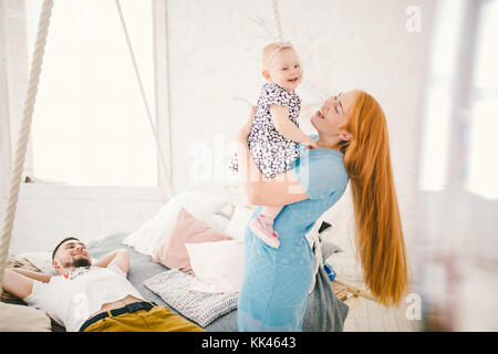 Belle jeune femme aux longs cheveux rouges dans une robe bleue est titulaire d'un enfant sur ses mains un an blonde près du lit sur lequel se trouve un homme au repos. Banque D'Images