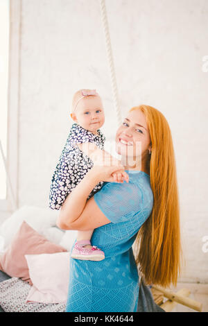 Belle jeune femme aux longs cheveux rouges dans une robe bleue est titulaire d'un enfant sur ses mains un an blonde près du lit sur lequel se trouve un homme au repos. Banque D'Images