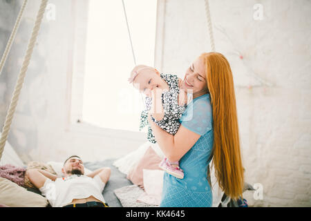 Belle jeune femme aux longs cheveux rouges dans une robe bleue est titulaire d'un enfant sur ses mains un an blonde près du lit sur lequel se trouve un homme au repos. Banque D'Images