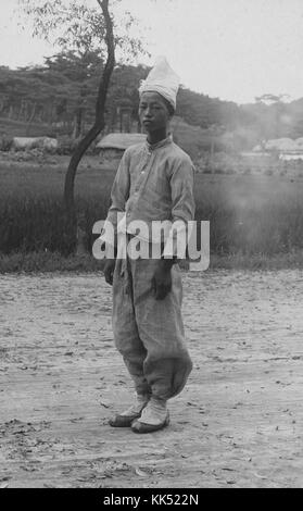 Portrait photographique en noir et blanc d'un coréen vêtu de vêtements paysans traditionnels, l'image a été capturée pendant l'occupation japonaise de la Corée, sa chemise et son pantalon sont faits du même matériau léger tandis que ses chaussures semblent tissées, un petit village peut être vu entre le champ herbeux et la forêt derrière lui, 1921. De la Bibliothèque publique de New York. Banque D'Images