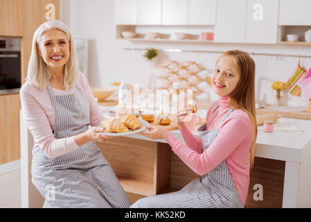 Le temps de cuisson. belle grand-mère et petite-fille positive heureux assis dans la cuisine et vous regarde tout en présentant leur nourriture Banque D'Images