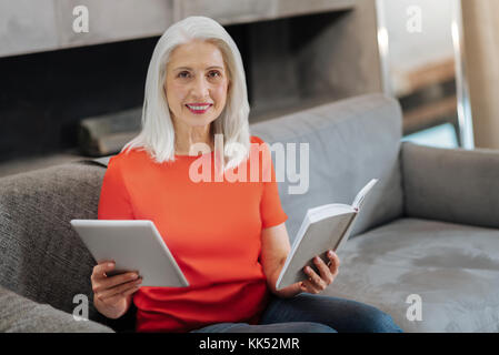 Dame agréable. belle femme âgée positive en vous regardant et souriant tout en comparant un livre papier aux technologies numériques Banque D'Images