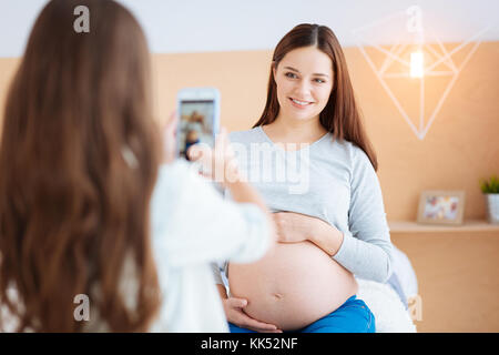 Moment heureux. belle jeune femme enceinte en souriant et serrant son ventre tout en fille taking photo Banque D'Images