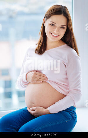 Mon amour. Portrait de femme enceinte optimiste aimant qui regarde droit serrant son ventre et posant contre la fenêtre Banque D'Images