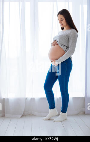 Mon soleil. femme enceinte attrayante positive article contre le tulle blanc tout en examinant son ventre et caressant Banque D'Images