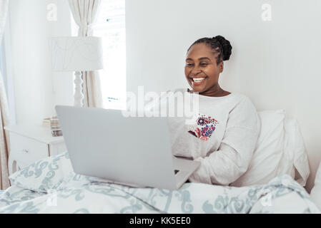 Souriante jeune femme africaine en utilisant un ordinateur portable dans son lit Banque D'Images