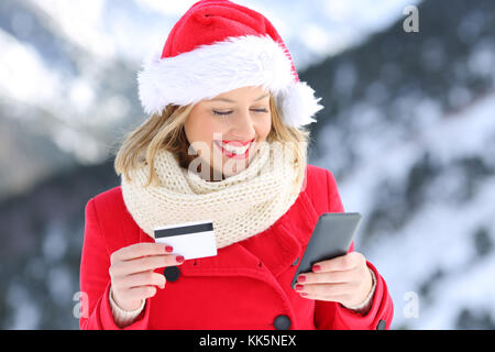 Happy woman wearing santa claus hat payer en ligne avec carte de crédit à Noël avec la montagne enneigée en arrière-plan Banque D'Images