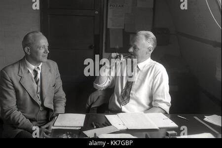 Années 1950, photo historique montrant un homme assis dans sa chemise et cravate à son bureau dans un bureau de tabac Fumer une pipe et d'avoir une discussion avec un autre homme. Pippe fumeurs était populaire chez les hommes adultes en Grande-Bretagne à cette époque. Banque D'Images