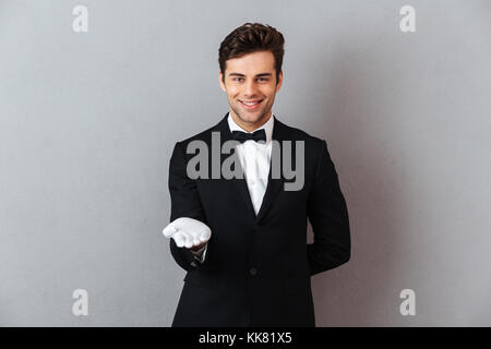 Portrait of a happy smiling homme habillé en smoking et permanent avec outsretched gants isolés à la main sur fond gris Banque D'Images