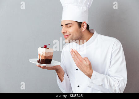 Portrait d'un homme habillé en chef satisfait odeur uniforme du gâteau sur une plaque sur fond gris isolé Banque D'Images