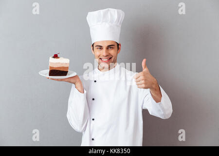 Portrait of a cheerful male chef habillés en uniforme de la plaque avec du gâteau et showing Thumbs up isolé sur fond gris des gestes Banque D'Images