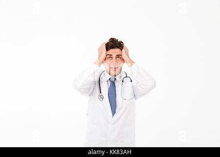 Portrait d'un jeune homme inquiet confus doctor with stethoscope habillés en uniforme debout, les bras à sa tête et à la recherche jusqu'isolated over white Banque D'Images