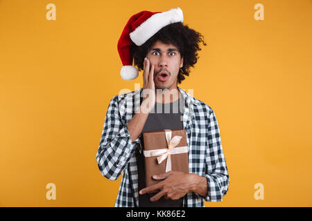 Portrait d'un choqué happy african man dressed in christmas hat holding gift box et à la caméra au plus isolé sur fond orange Banque D'Images