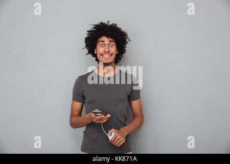 Portrait of a smiling casual african man holding mobile phone connecté à l'alimentation sur fond gris isolé banque Banque D'Images