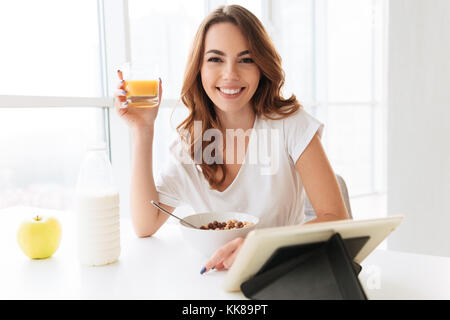 Image of dame assise à la cuisine près de la fenêtre. Appareil photo à manger des corn flakes boire du jus à l'aide de l'ordinateur tablette. Banque D'Images