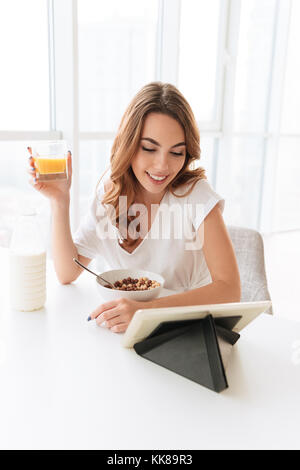 Image femme assise à la cuisine près de la fenêtre. À côté des flocons de maïs manger boire du jus à l'aide de l'ordinateur tablette. Banque D'Images