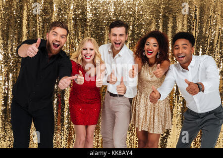 Groupe d'amis tout en fête multiculturelle joyeuse et permanent showing Thumbs up plus isolé geste brillant or background Banque D'Images